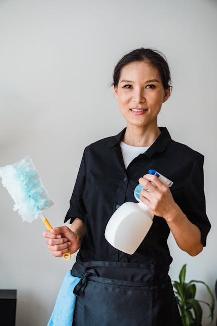 A professional cleaner from Barnes Carpet Cleaning stands indoors against a plain, light-colored wall, holding a spray bottle of cleaning solution in one hand and a cleaning cloth in the other. The individual is dressed in a black cleaning uniform with a white shirt underneath, and has a friendly expression. In the background, there is a partial view of a contemporary kitchen with a black countertop and a potted plant. The surface area in the image appears tidy and well-maintained, reflecting thorough cleaning and hygiene practices typical of domestic or commercial surface cleaning services for floors, countertops, or other surfaces, emphasizing cleanliness and sanitisation properties. Barnes Carpet Cleaning specializes in deep cleaning and sanitisation, ensuring surfaces are spotless, shiny, and hygienic.