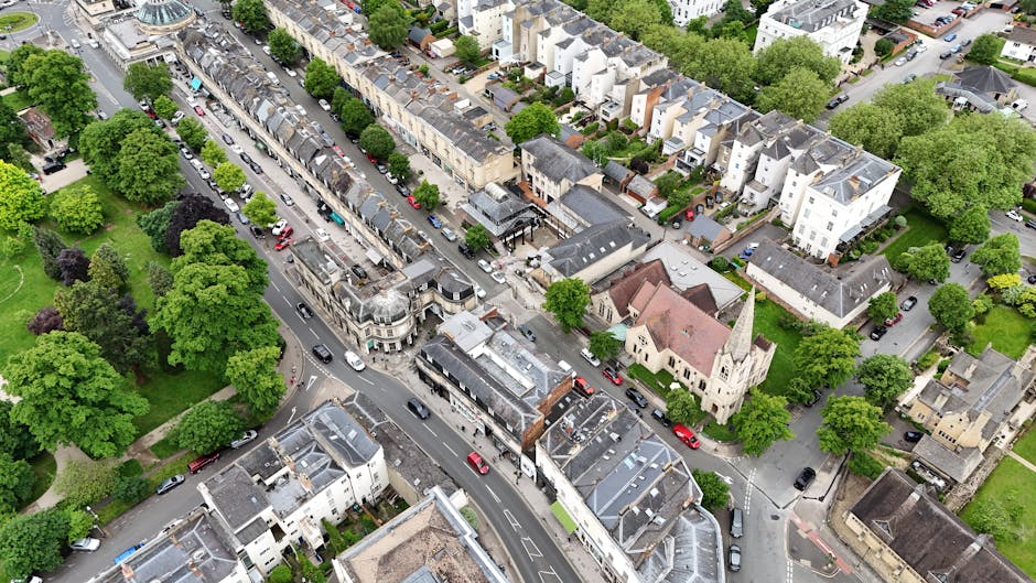 An aerial view of Barnes High Street in SW13, showing a busy urban street lined with shops, cafes, and parked cars. The street features a mix of traditional and modern buildings, including a notable church with a tall steeple and a curved facade. Surrounding the street are residential houses with pitched roofs, some with rooftop terraces, amid lush green trees that line the sidewalks and fill the neighboring parks. The scene is well-lit with natural daylight, highlighting the cleanliness and orderliness of the area. Barnes Carpet Cleaning specializes in surface cleaning and deep cleaning services for local homes and businesses, ensuring thorough sanitisation and maintenance of commercial and domestic interiors.