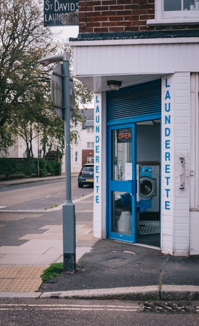 The image shows a small laundry depot positioned on a street corner, featuring a white wooden exterior with vertical blue signage reading 'LAUNDERETTE' on either side of the entrance. The glass door reveals a blue washing machine inside, with an illuminated 'OPEN' sign hung above. In front of the entrance, there's a metallic street lamp and a vending machine or possibly an advertisement display attached to a pole. The sidewalk is clean, with a tactile paving strip for accessibility, and a parked car is visible further down the street. Behind the laundry depot, trees and other buildings are visible under a cloudy sky. Barnes Carpet Cleaning's name is not visible in this image, but this scene represents a typical street-side utility facility emphasizing cleanliness and maintenance in a residential or commercial area. It reflects the importance of surface cleaning and hygiene for keeping such spaces tidy and operational, aligning with the themes of deep cleaning and sanitisation associated with the Barnes High Street carpet cleaning guide SW13, BARNES.