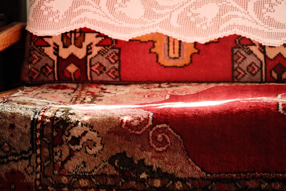Close-up of a vintage patterned upholstered sofa with a red and beige Persian-style rug on the armrest, illuminated by warm natural light. A white lace doily partially covers the top of the sofa, which appears clean and well-maintained, highlighting the intricate design and vibrant colors of the textiles. The setting suggests a cozy, traditionally decorated living room, emphasizing surface cleaning and upholstery care. Barnes Carpet Cleaning specializes in deep cleaning and sanitisation for domestic furniture, ensuring hygiene and preservation of delicate materials. The scene reflects careful surface maintenance typical of professional cleaning services offered by Barnes Carpet Cleaning, as shown on the page about Lonsdale Road residents rug and upholstery cleaning tips.