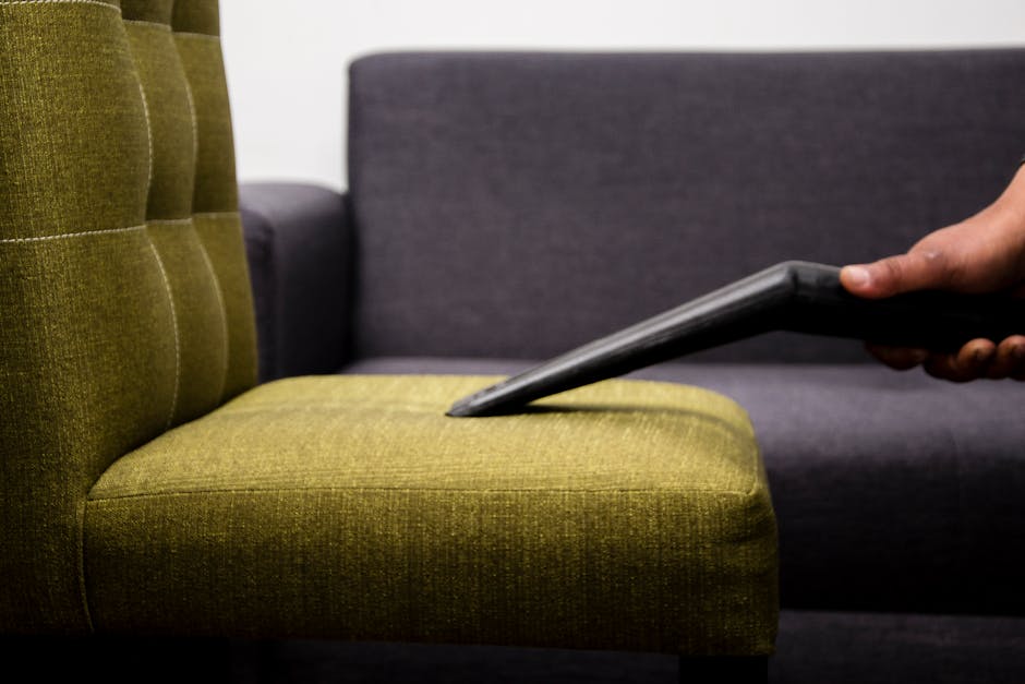 Close-up of a person using a handheld vacuum cleaner to surface clean a green upholstered armchair, with other similar modern chairs in grey fabric visible in the background, in an indoor setting with soft lighting, demonstrating furniture deep cleaning and upholstery hygiene as part of domestic cleaning services provided by Barnes Carpet Cleaning.